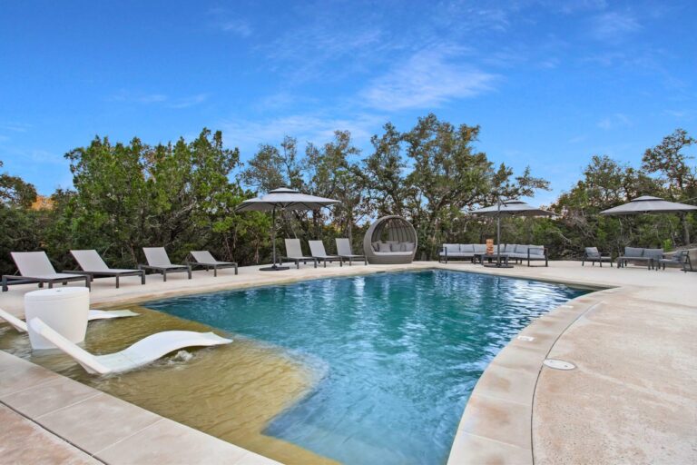 Rectangular pool at Starlight Horizon with lounge chairs, umbrellas, and a curved wicker daybed along the deck, framed by Hill Country trees