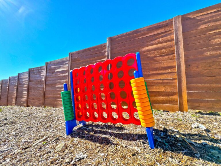 Giant red Connect Four lawn game with green and yellow chip stacks set up on mulch in front of the wooden privacy fence at Starlight Horizon